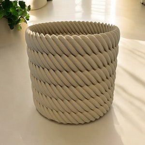 May include: A beige woven planter with a rope-like design. The planter is made of a durable material and has a smooth finish.