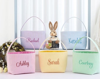 May include: Five pastel-colored Easter baskets with embroidered names. Each basket has a seersucker stripe pattern and a contrasting interior lining. A decorative bunny figurine is placed between the baskets.