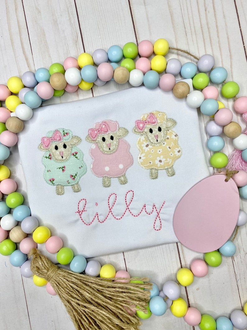 May include: White shirt with three embroidered sheep, each with a different floral pattern and pink bow. The name "Tilly" is stitched in pink below the sheep. A pastel bead garland and pink egg-shaped tag surround the shirt.