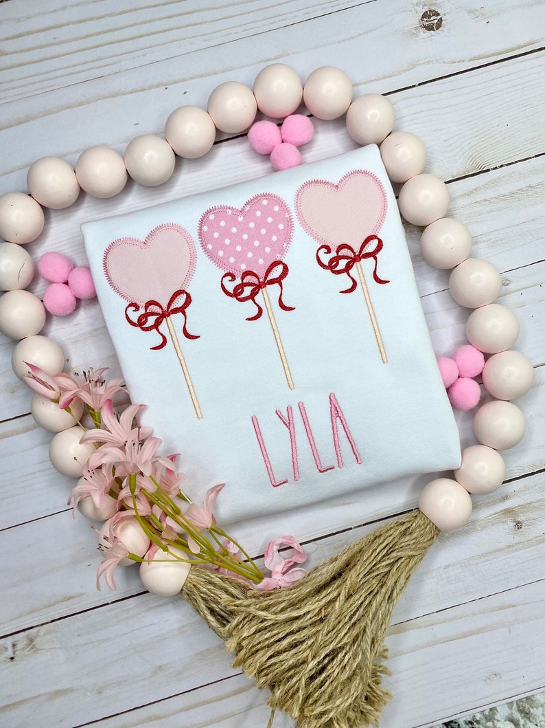 Op de afbeelding: Wit T-shirt met drie geborduurde hartvormige lolly's, elk met een rode strik. E&eacute;n hart heeft een roze stippenpatroon. De naam "LYLA" is roze geborduurd. Het T-shirt is versierd met roze bloemen en houten kralen.
