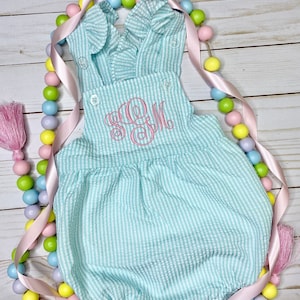 May include: A seersucker romper in a light blue and white striped pattern, featuring a ruffled neckline and straps. The romper has a pink embroidered monogram and is adorned with decorative buttons. A pink ribbon and pastel-colored beads surround the romper.