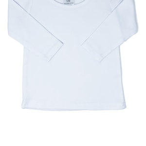 May include: A white long-sleeved shirt with a round neckline and a tag that reads "AJ Blanks 12M". The shirt has a simple design with a slightly puffed sleeve detail at the shoulder. The garment is made of 100% cotton.