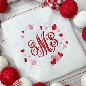 May include: White square shirt with a red and pink heart design. The design features a monogram with the letters "MS" surrounded by a heart-shaped wreath. Red and white decorative balls surround the shirt.
