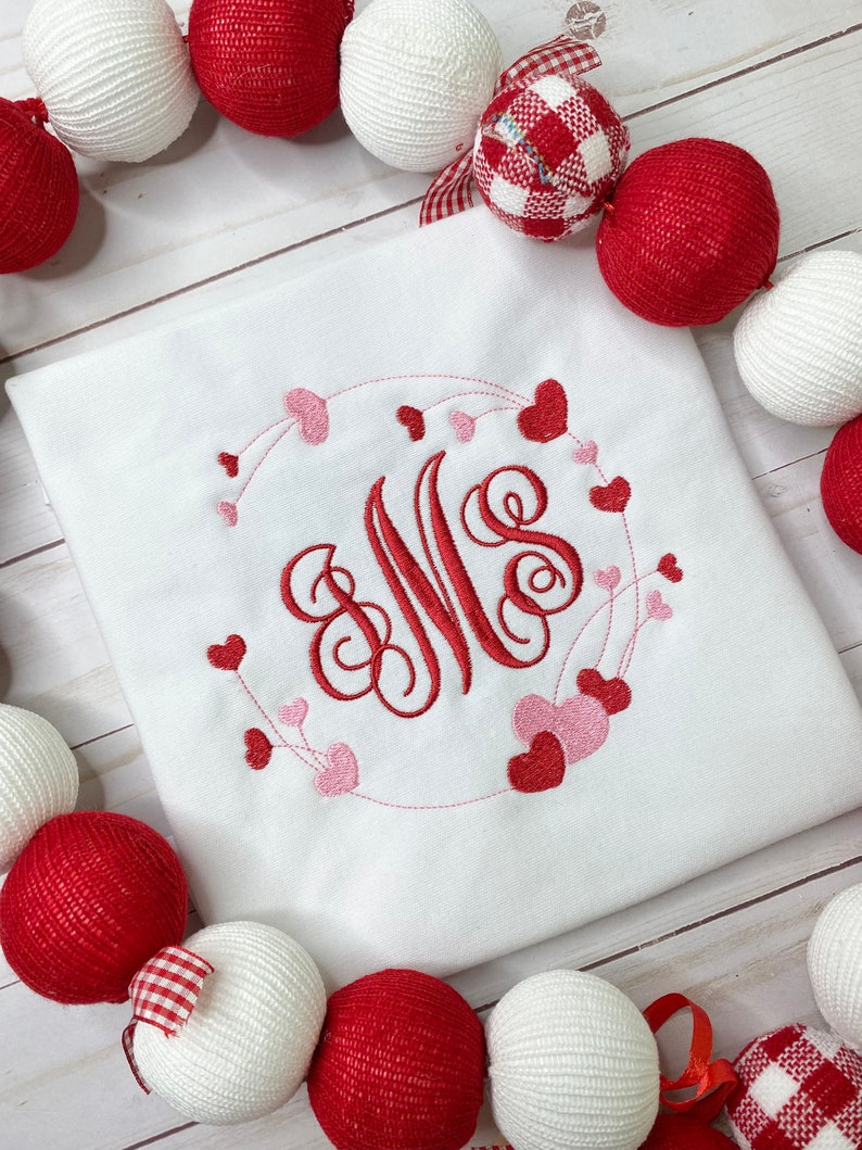 May include: White square shirt with a red and pink heart design. The design features a monogram with the letters "MS" surrounded by a heart-shaped wreath. Red and white decorative balls surround the shirt.