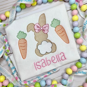 May include: White Easter-themed shirt with a bunny design, pink bow, and carrots. The name "Isabella" is embroidered in pink. Decorative pastel-colored beads and a ribbon with "Happy Easter" text surround the shirt.
