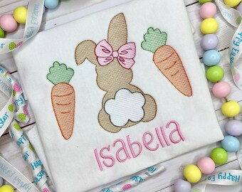 May include: White Easter-themed shirt with a bunny design, pink bow, and carrots. The name "Isabella" is embroidered in pink. Decorative pastel-colored beads and a ribbon with "Happy Easter" text surround the shirt.