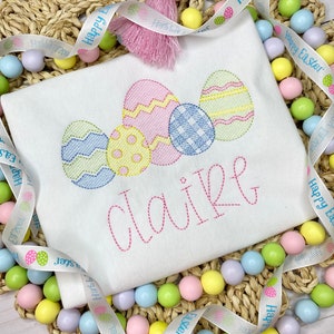 May include: White t-shirt embroidered with colorful Easter eggs and the name "Claire" in pink script. The shirt is decorated with a pink tassel and surrounded by pastel-colored beads and a ribbon that says "Happy Easter".