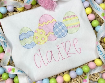 May include: White t-shirt embroidered with colorful Easter eggs and the name "Claire" in pink script. The shirt is decorated with a pink tassel and surrounded by pastel-colored beads and a ribbon that says "Happy Easter".