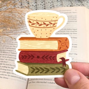 May include: A die-cut sticker featuring a stack of three books with a cream-colored teacup resting on top. The top book is orange, the middle book is red with a dark bookmark, and the bottom book is green. Each book spine features a decorative leaf pattern.