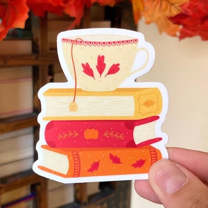 Peut inclure: Un autocollant représentant une pile de trois livres surmontée d'une tasse à thé. Les livres sont orange, rouge et jaune, décorés de feuilles et d'une citrouille. La tasse à thé est crème avec des motifs de feuilles rouges.