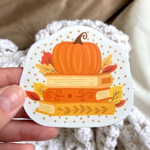 May include: A die-cut sticker featuring a stack of three books in shades of yellow and orange, topped with a bright orange pumpkin. The design is accented with colorful autumn leaves in red, orange, and yellow, set against a background of small dots.