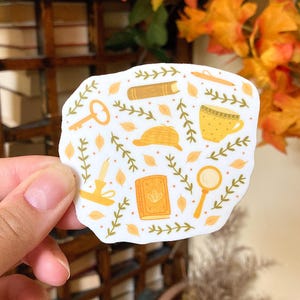 May include: A white sticker with illustrations of a key, book, magnifying glass, candle, hat, and teacup. The sticker has a detective theme with a yellow and brown color scheme, surrounded by green leaf accents.