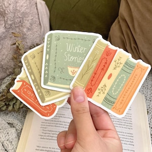 May include: A hand holds several book-shaped stickers. The stickers feature illustrations of books in various colors, including green, orange, and beige. One sticker reads "Winter Stories". The stickers are on a book and a green and beige background.