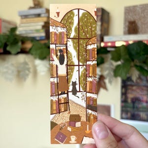 May include: A rectangular paper bookmark featuring an illustration of a person on a ladder reaching for books on shelves flanking a large arched window. A black cat sits on the floor near the window, and a table with books and a cup sits below.