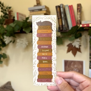 Peut inclure: Un marque-page en papier rectangulaire présentant une pile verticale de livres illustrés dans des tons marron, rouge et doré. Un chat gris repose sur le livre du haut. Le texte indique "EVERYDAY is a good DAY TO read a BOOK". Une bordure de vigne délicate encadre la pile.