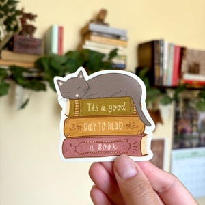 May include: A die-cut sticker featuring a gray cat sleeping on a stack of three books. The top book is olive green with the text 'It's a good', the middle book is yellow with 'DAY TO READ', and the bottom book is maroon with 'a BOOK'.