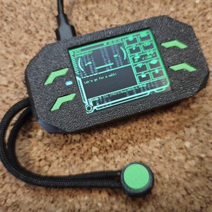 Könnte beinhalten: Schwarzes Handheld-Gerät mit grünem Bildschirm, der Text und Grafiken anzeigt. Das Gerät hat grüne Akzente und einen grünen Knopf an einem schwarzen Kabel. Der Bildschirm zeigt "Let's go for a walk!"