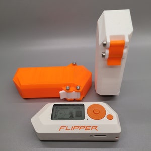 Könnte beinhalten: Ein weiß-oranges Flipper-Gerät mit Bildschirm, Knöpfen und dem Aufdruck "FLIPPER". Zwei orangefarbene und weiße Gehäuse sind ebenfalls vorhanden, eines rechteckig und eines hoch, mit Metallscharnieren. Die Gegenstände befinden sich auf einer grauen Oberfläche.