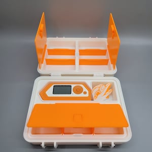 May include: An orange and white plastic storage case with a small orange device and a white object inside. The case has a hinged lid and multiple compartments.