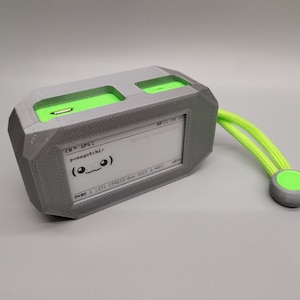May include: A gray 3D printed device with a green strap and a small green button. The device has a small screen that displays the text "CH * AP51 pmayatchi> (o_o)" and "IP 01 04 20".