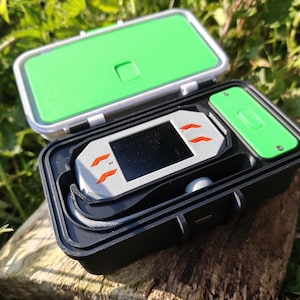 May include: A black, rectangular case with a green lid and a white and green interior. Inside, a silver device with an orange design and a black screen rests, along with a small green accessory. The case is open, revealing the contents.