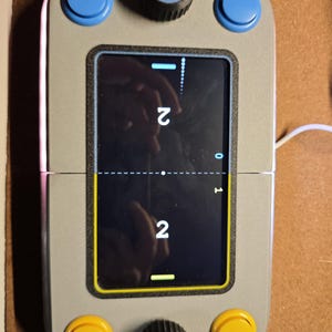 Pode incluir: Um dispositivo de jogo portátil retangular cinza com uma tela central exibindo um jogo de pong digital com pontuações de 2 e 2. O dispositivo inclui dois botões giratórios pretos, dois botões circulares azuis no topo e dois botões circulares amarelos na base.