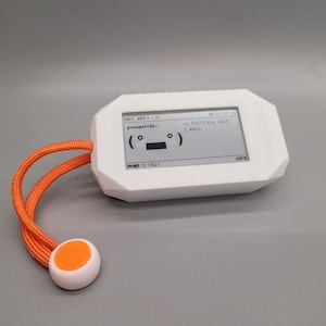 Op de afbeelding: Een wit, geometrisch gevormd elektronisch apparaat met een klein scherm dat tekst en een eenvoudige afbeelding weergeeft. Het heeft een oranje koord met een witte en oranje bal aan het uiteinde. Het scherm toont de tekst "p-nagotchi" en "To FRITZ!Box 0515 2,4 GHz".