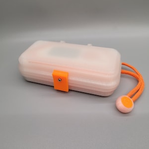 Könnte beinhalten: Eine weiße Plastik-Aufbewahrungsbox mit einem orangefarbenen Verschluss und einer passenden orangefarbenen Schnur mit einem runden orangefarbenen Ende. Die Box hat eine strukturierte Oberfläche.