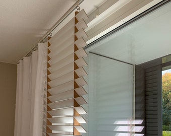 May include: White wooden blinds with a light brown edge are installed next to a large window. Sheer white curtains hang to the left of the blinds. The blinds are partially open, allowing light to filter through.