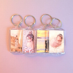 May include: Four clear acrylic keychains with gold-colored rings. Each keychain holds a different photograph. The photos include a couple on a cliff, a woman with her hair up, a silhouette of a couple at sunset, and a newborn baby wrapped in a blanket.