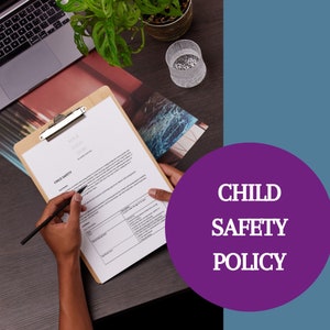 May include: A person reviewing a Child Safety Policy document on a clipboard. The document has a section titled "Child Safety" and includes a table with information about safety procedures. The background is a wooden table with a plant and a glass object.
