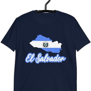May include: Navy blue t-shirt featuring a graphic of the El Salvador flag within the country's outline. The text "El Salvador" is written in a light blue script below the graphic. The shirt is hanging on a black hanger.