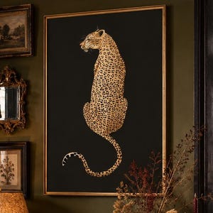 May include: A framed wall art print featuring a gold leopard illustration on a black background, displayed above a dark wood console table with a gold lamp, a leopard print vase with dried branches, and a stack of books in a room with dark walls.