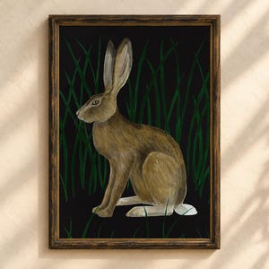 May include: A framed wall art print featuring a brown hare sitting in tall green grass against a solid black background. The hare is depicted in profile, facing left, with long ears and a white tail. The frame is a dark, textured wood finish.