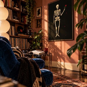 May include: A large rectangular wall art piece featuring a white human skeleton on a black background, holding a single red rose. The artwork is framed in a thin gold border and mounted on a dusty pink wall in a room with a blue velvet chair.