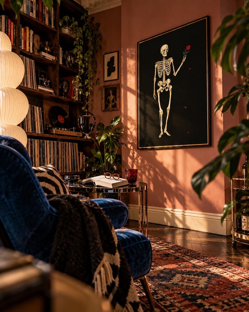 May include: A large rectangular wall art piece featuring a white human skeleton on a black background, holding a single red rose. The artwork is framed in a thin gold border and mounted on a dusty pink wall in a room with a blue velvet chair.