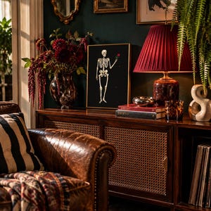 May include: A framed wall art print featuring a white skeleton on a black background, holding a single red rose. The artwork is displayed on a wooden sideboard next to a dark red pleated lampshade, a vase of dark flowers, and books.