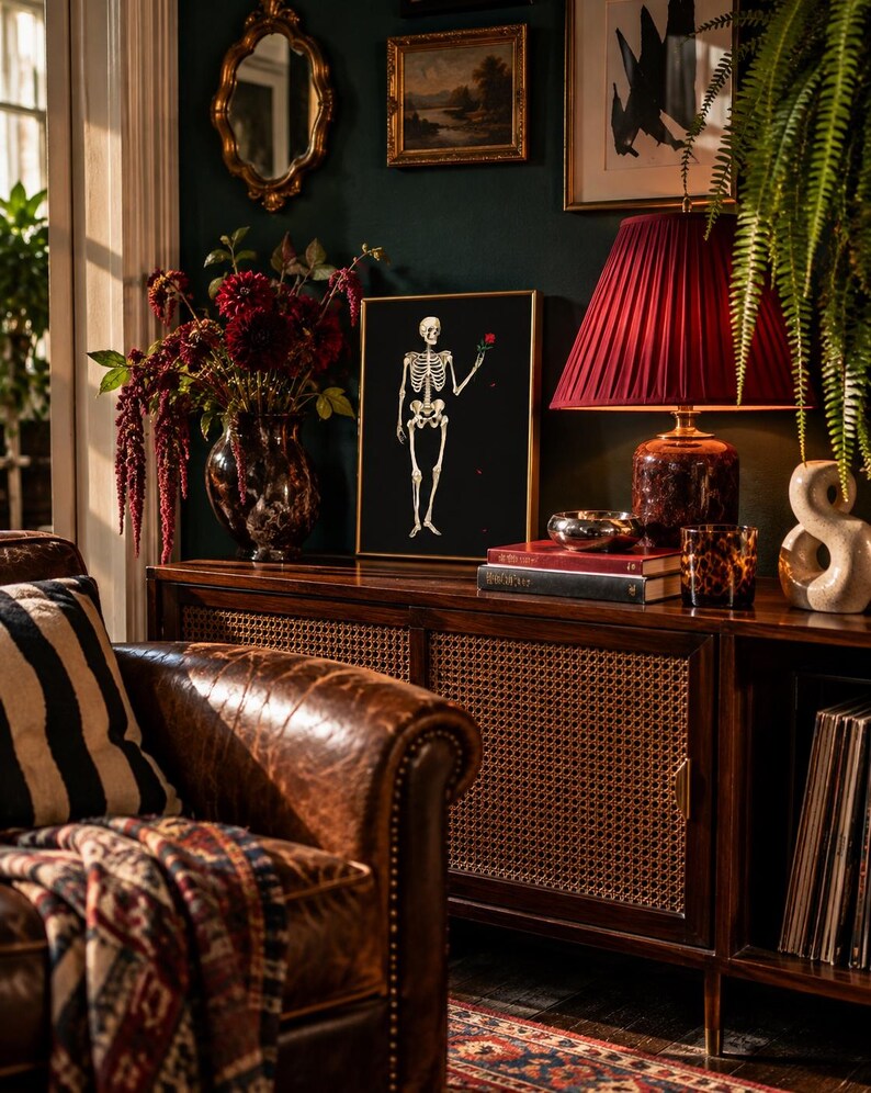 May include: A framed wall art print featuring a white skeleton on a black background, holding a single red rose. The artwork is displayed on a wooden sideboard next to a dark red pleated lampshade, a vase of dark flowers, and books.