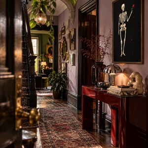 May include: A hallway interior featuring a glossy dark red console table, a framed black wall art piece depicting a skeleton holding a red rose, a patterned runner rug, a dark staircase with leopard print carpet, and various decorative home accents.