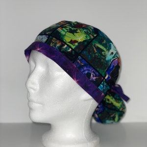 May include: A patterned scrub cap featuring a colorful design with green, blue, and purple hues. The cap has a dark purple band with a starry pattern and a matching tie in the back. The cap is displayed on a white mannequin head.