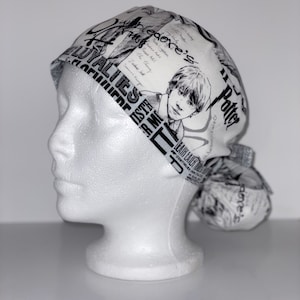 May include: A white and black patterned scrub cap featuring a portrait of a young man and text from a newspaper. The cap is tied in a bun at the back. The cap is on a white mannequin head.