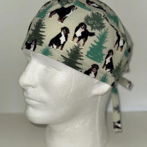 May include: A light green surgical scrub cap featuring a repeating pattern of Bernese Mountain Dogs and green pine trees. The cap has a tie closure at the back and is displayed on a white mannequin head.