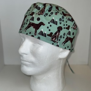 May include: A light green surgical scrub cap featuring a repeating pattern of brown dogs, black paw prints, mountains, and footprints. The cap is tied at the back and is displayed on a white mannequin head.