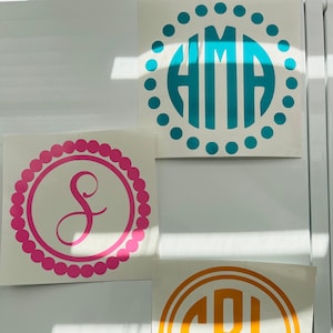 May include: Three white squares with different colored monogram decals. The top decal is turquoise blue with the letters "AMA" in a circle. The middle decal is pink with the letter "S" in a circle. The bottom decal is orange with the letters "SBL" in a circle.