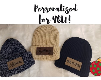 May include: Three personalized knit beanies with leather patches. The beanies are beige, blue, and gray. The patches have the names "Sydney", "Lillianna", and "Oliver" written on them. A red and green Christmas ornament is in the background.