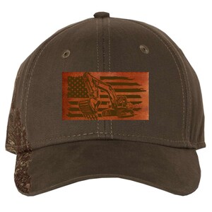 Op de afbeelding: Een bruine baseballpet met een bruine leren patch met een excavator-silhouet tegen een Amerikaanse vlagontwerp.