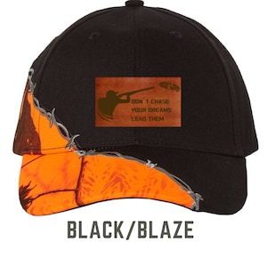 May include: Black baseball cap with an orange blaze camouflage brim. The cap has a brown leather patch with the text "Don't chase your dreams, lead them".