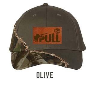 Może przedstawiać: Zielona bejsbolówka z daszkiem w kamuflażu i brązową skórzaną naszywką z napisem "PULL" i sylwetką osoby trzymającej strzelbę.