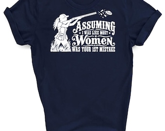 May include: A navy blue short-sleeved t-shirt featuring a white graphic of a person shooting a clay target with a shotgun. The text reads: Assuming I was like most women was your 1st mistake. The design includes a silhouette of a shooter in tall grass.