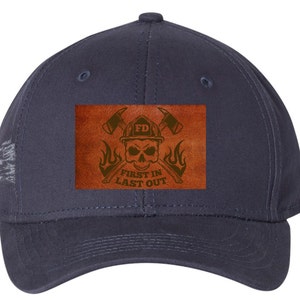 Könnte beinhalten: Eine marineblaue Baseballkappe mit einem braunen Lederpatch, auf dem ein Schädel mit gekreuzten Äxten, Flammen und dem Text "FD FIRST IN LAST OUT" abgebildet ist.